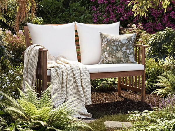أريكة خشبية خارجية بمقعدين مزودة بوسائد بيضاء، ووسادة بنقشة زهور، وبطانية مزخرفة، في حديقة غنية بالنباتات السرخسية والأزهار المتفتحة. اضغط لمشاهدة المنتجات في المجموعة.
